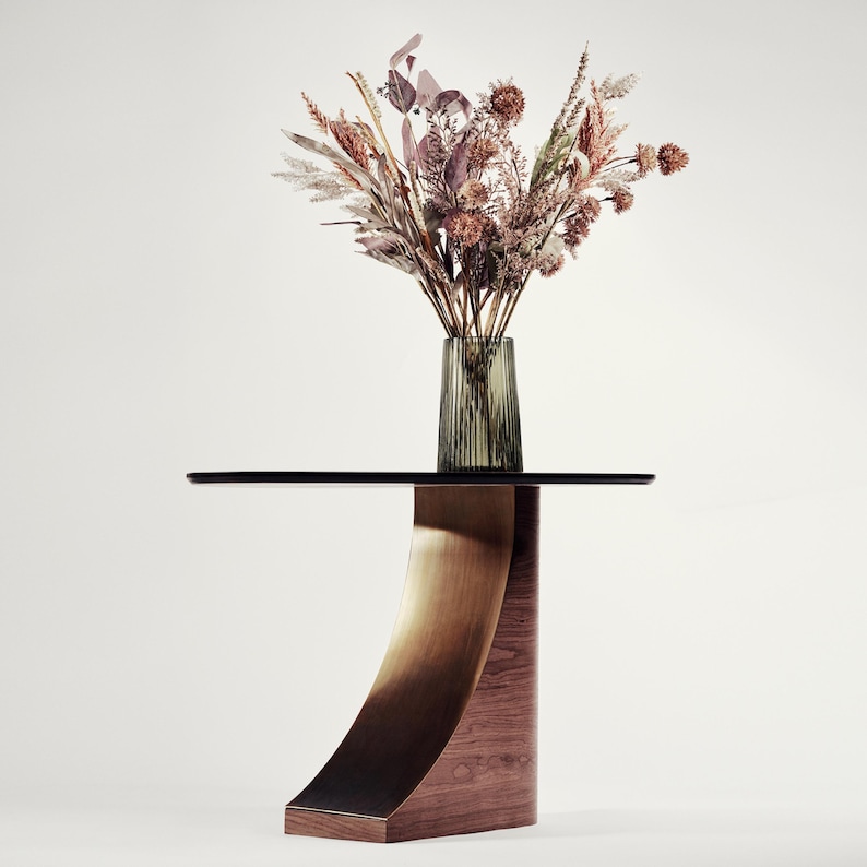 May include: A modern side table with a black glass top and a curved, bronze-toned metal support. The base is made of wood. A vase of dried flowers in shades of brown, beige, and purple sits on the table.