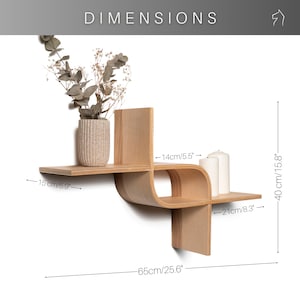 May include: A light-colored wooden wall shelf with a unique, curved design. The shelf is 25.6 inches wide and 15.8 inches high. It features two shelves and a vertical support. A vase with dried flowers and two white candles are displayed on the shelf.