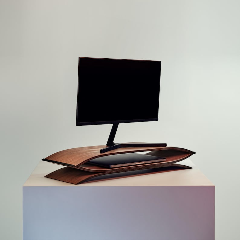 May include: A dark wood monitor stand featuring two curved, layered shelves with a smooth finish. The top shelf supports a black computer monitor with a thin stand, while the bottom shelf provides storage space for a closed laptop computer.
