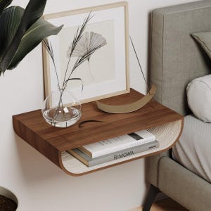 Puede incluir: Una estantería flotante de madera con un borde curvo, montada en una pared blanca. La estantería está decorada con un jarrón de vidrio lleno de ramas secas, una escultura de metal dorado y una pila de libros con el título "RIMOWA" visible. La estantería está al lado de una cama con una cabecera gris claro.