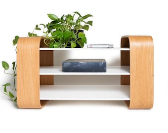 May include: A low-profile shelving unit featuring two curved light-toned wood side panels and three horizontal white shelves. A green leafy plant sits on the top shelf, and a dark rectangular book is placed on the middle shelf.