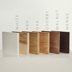 May include: Six rectangular wood samples arranged in a row, labeled White, Ash, Oak, Cherry, Walnut, and Wenge. The samples display varying natural wood grain patterns and colors, ranging from a solid white finish to light tan, medium brown, and dark brown.