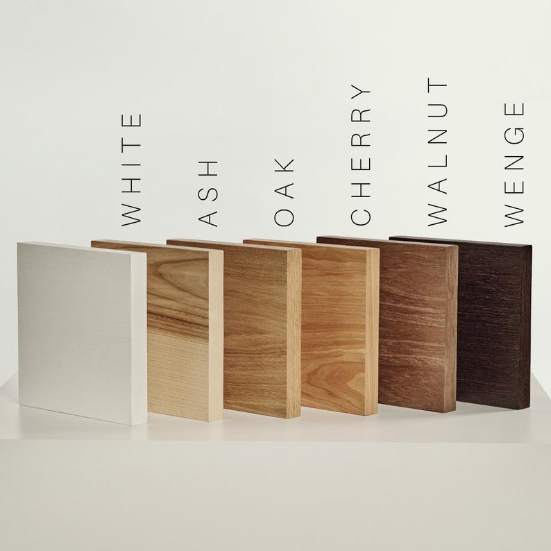 May include: Six rectangular wood samples arranged in a row, labeled White, Ash, Oak, Cherry, Walnut, and Wenge. The samples display varying natural wood grain patterns and colors, ranging from a solid white finish to light tan, medium brown, and dark brown.
