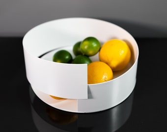 May include: A white, modern fruit bowl with a circular design. The bowl contains a mix of fresh fruit, including bright yellow lemons and green limes. The bowl is sitting on a reflective black surface.