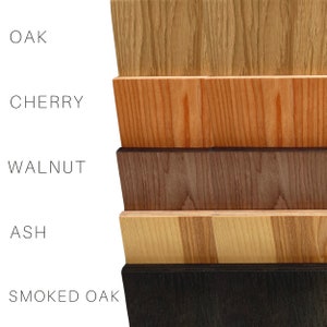Op de afbeelding: Een close-up van zes houten stalen, elk met een ander nerfpatroon en kleur. De stalen zijn gelabeld met "OAK", "CHERRY", "WALNUT", "ASH" en "SMOKED OAK". De houtkleuren vari&euml;ren van lichtbruin tot donkerbruin en zwart.