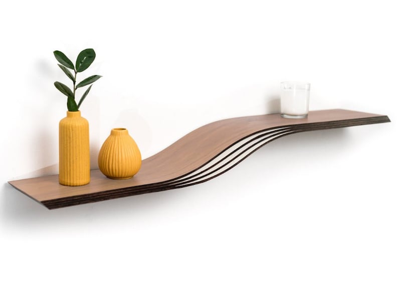 May include: A modern, floating wall shelf with a unique wave design. The shelf is made of layered wood and holds two yellow vases with plants and a small candle in a glass. The shelf is mounted on a white wall.