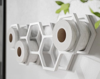 May include: White honeycomb-shaped toilet paper holder mounted on a white wall. The holder is designed to hold multiple rolls of toilet paper, with the rolls visible in the hexagonal openings. The design is modern and minimalist.