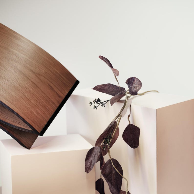 May include: A curved wooden object with a rich brown grain and black edges, positioned next to a delicate stem of dark purple eucalyptus leaves. The wooden piece features a smooth, polished surface and a distinct, angled, layered construction.