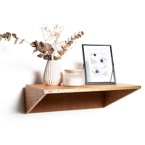 May include: A wooden floating shelf with a vase of dried flowers, a framed print with eye illustrations, and a candle in a glass jar. The shelf has a natural wood grain finish and a unique angled design.