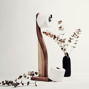 May include: A wooden toilet paper holder with a curved design, featuring a roll of white toilet paper. A second roll of toilet paper rests on the surface. A black vase holds dried branches. The holder is made of dark and light wood.