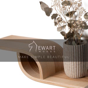 Pu&ograve; includere: Una moderna mensola in legno dal design curvo, con un vaso in ceramica grigio e beige con fiori secchi. La mensola presenta il testo "EWART WOODS" e "MAKE SIMPLE BEAUTIFUL".