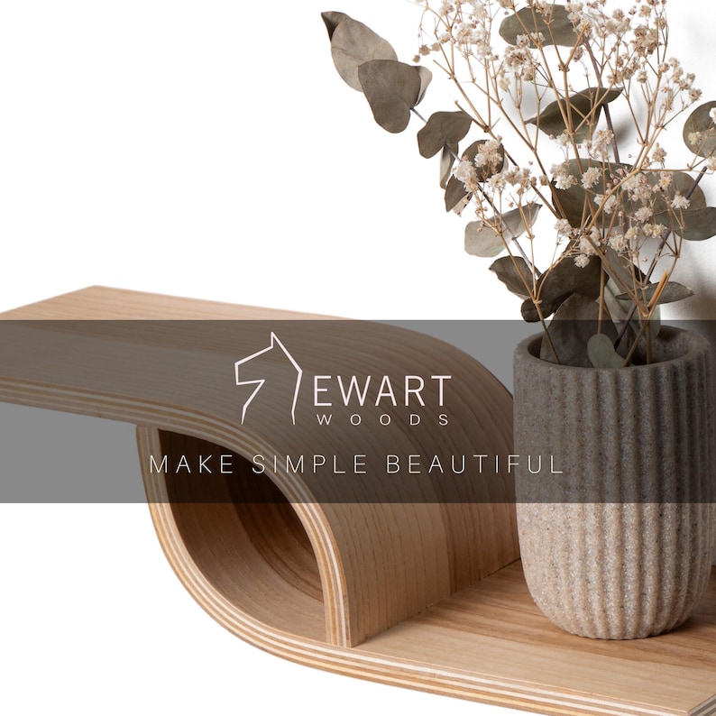 Pu&ograve; includere: Una moderna mensola in legno dal design curvo, con un vaso in ceramica grigio e beige con fiori secchi. La mensola presenta il testo "EWART WOODS" e "MAKE SIMPLE BEAUTIFUL".