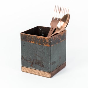 Square utensil holder with a rustic copper finish and wooden base, containing copper-colored forks and a spoon. The holder is placed against a plain white background.