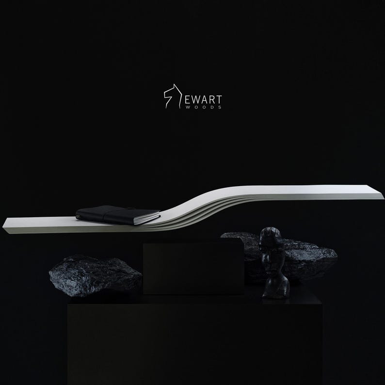 May include: A minimalist, modern display featuring a white, curved shelf with a black notebook. Below, a black box and a small, dark sculpture are displayed with dark rocks against a black background. The "Ewart Woods" logo is visible.