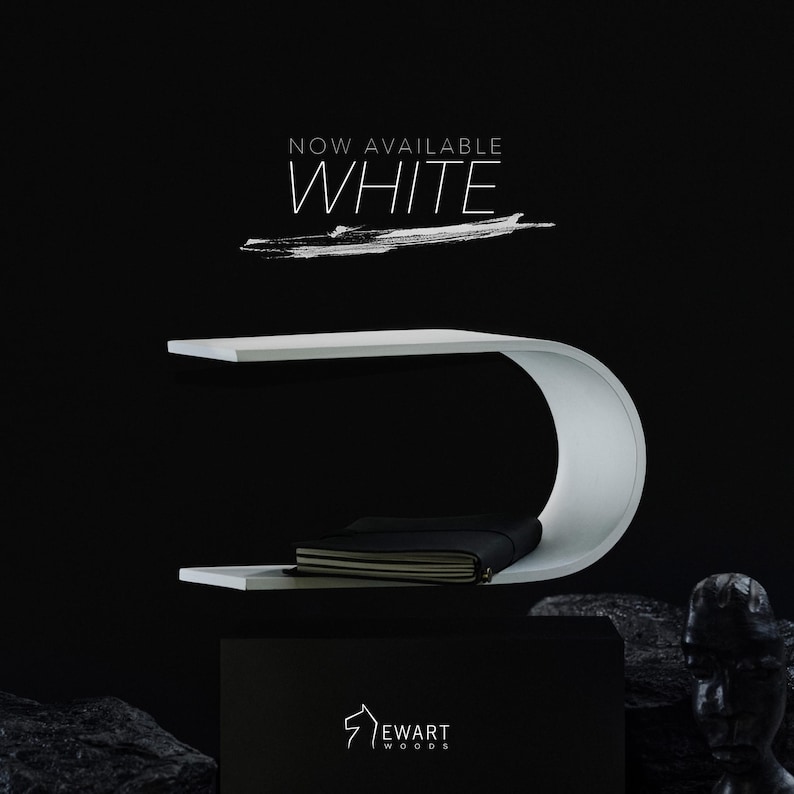 May include: A modern white floating shelf with a curved design, holding a black notebook. The text "NOW AVAILABLE WHITE" is displayed above the shelf. The brand logo "EWART WOODS" is at the bottom.