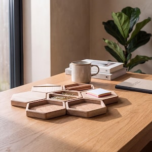 Peut inclure: Un ensemble d'organisateurs de bureau modulaire composé de six plateaux hexagonaux en bois imbriqués. Les plateaux présentent un grain de bois clair, avec deux unités contenant des inserts en liège, une contenant des trombones dorés, une des crayons.