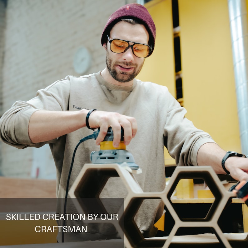 May include: A person wearing a maroon beanie and amber-tinted glasses uses a yellow and gray electric orbital sander to smooth the edges of a light-colored wooden hexagonal shelf unit. The text SKILLED CREATION BY OUR CRAFTSMAN is visible.