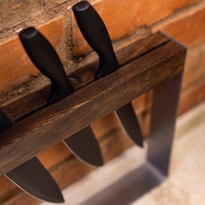 Top-down view of a knife holder with a dark wood block and sleek metal frame, holding two black-handled knives, positioned against a textured red brick kitchen wall.