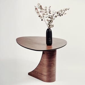 May include: A modern, wooden side table with a unique, curved base and a dark, oval tabletop. A black vase with a face design holds dried branches. The table's wood grain is visible, and the overall aesthetic is minimalist.