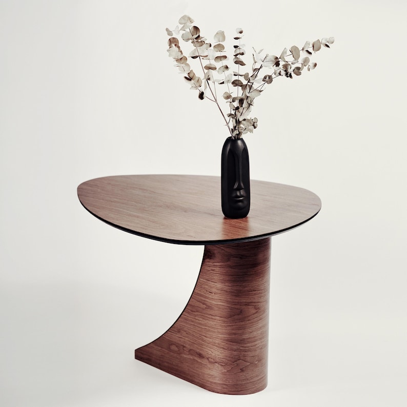 May include: A modern, wooden side table with a unique, curved base and a dark, oval tabletop. A black vase with a face design holds dried branches. The table's wood grain is visible, and the overall aesthetic is minimalist.
