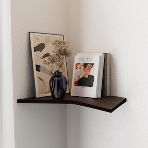 May include: A dark brown corner shelf with a curved edge, holding a framed print, a blue vase with dried flowers, and a stack of books with the title "SCHIELE" on the top book.