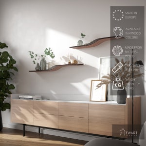 May include: Two modern, wavy wooden shelves in a living room setting. The shelves are a dark brown color with a layered design. The shelves are mounted on a white wall above a light wood cabinet. The image also shows decorative items on the shelves and cabinet. The text in the image says "MADE IN EUROPE", "AVAILABLE IN 4 WOOD COLORS", "MADE FROM NATURAL WOOD", "TWO SIZES AVAILABLE", and "WEIGHT CAPACITY ~5KG".