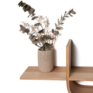 May include: A beige, textured vase with dried flowers and eucalyptus leaves sits on a light wood shelf. The shelf is part of a modern, minimalist wall-mounted shelving unit, showcasing a blend of natural elements and contemporary design.