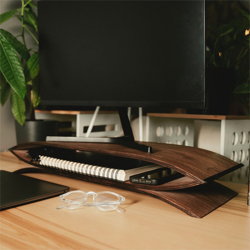 May include: A curved dark wood monitor stand with a hollow storage compartment holding a spiral-bound notebook and a smartphone. The stand features a sleek, layered design with a smooth finish, providing an elevated base for a computer monitor.