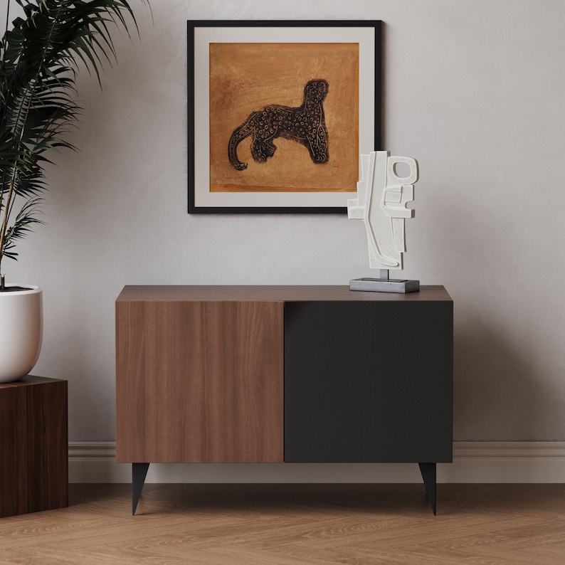 K&ouml;nnte beinhalten: Ein Holzschr&auml;nkchen mit einer schwarzen T&uuml;r und zwei schwarzen Beinen. Der Schrank steht auf einem Holzboden. &Uuml;ber dem Schrank h&auml;ngt ein gerahmtes Bild mit einer schwarzen Panther.