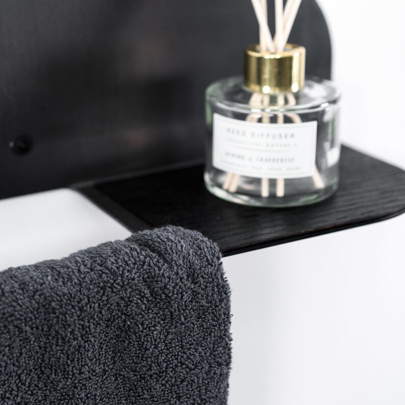 May include: A black shelf with a gray towel hanging from it. A clear glass bottle with a gold lid and a white label that says "Reed Diffuser" is on the shelf.