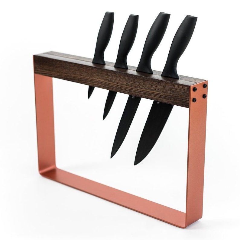 Angled view of a stylish knife stand with a dark wooden top, vibrant copper metal frame, and visible black screws, holding four black-handled knives in a single vertical slot.