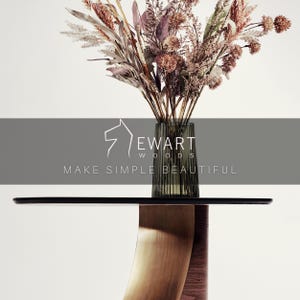 May include: A modern side table with a dark top and a curved wooden base. A clear glass vase with vertical lines holds a bouquet of dried flowers in shades of brown, pink, and beige. The text "EWART WOODS MAKE SIMPLE BEAUTIFUL" is displayed.