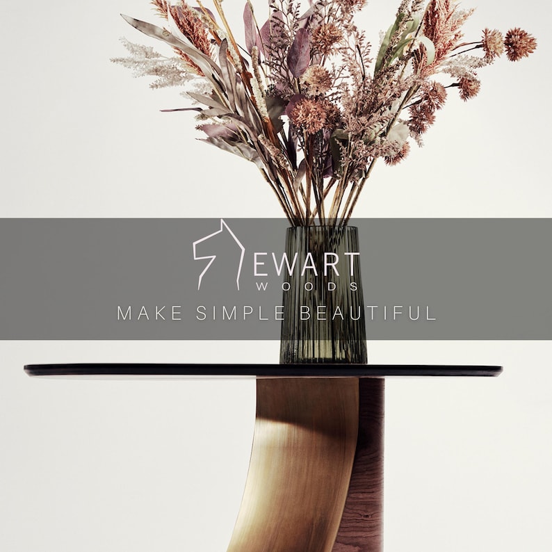 May include: A modern side table with a dark top and a curved wooden base. A clear glass vase with vertical lines holds a bouquet of dried flowers in shades of brown, pink, and beige. The text "EWART WOODS MAKE SIMPLE BEAUTIFUL" is displayed.