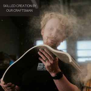 May include: A craftsman holding a curved wooden piece, with sawdust in the air. The text "SKILLED CREATION BY OUR CRAFTSMAN" is at the top left. The craftsman is wearing a black t-shirt with white text.