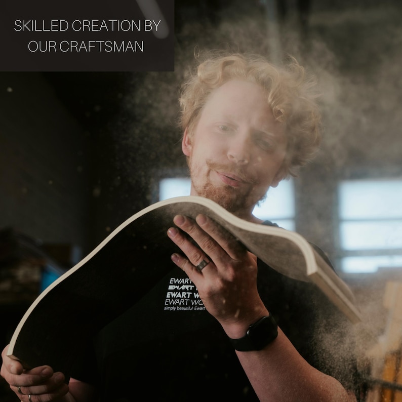May include: A craftsman holding a curved wooden piece, with sawdust in the air. The text "SKILLED CREATION BY OUR CRAFTSMAN" is at the top left. The craftsman is wearing a black t-shirt with white text.