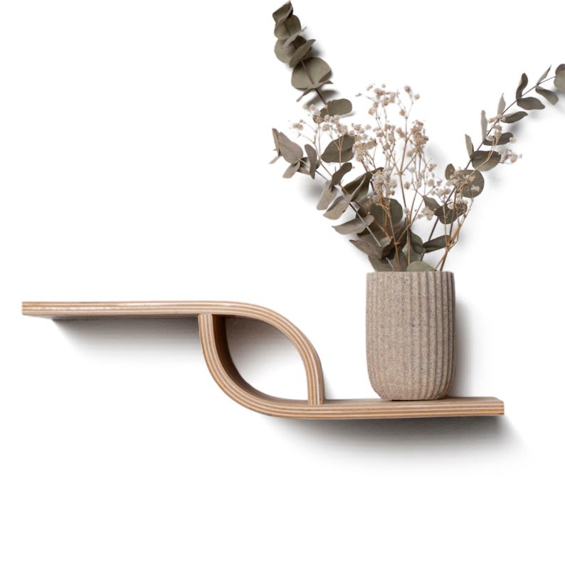 Pu&ograve; includere: Una moderna mensola da parete in legno chiaro, con un design unico a forma di foglia. Un piccolo vaso beige strutturato contiene fiori secchi e verde. La mensola &egrave; montata su una parete bianca, creando un'estetica minimalista.