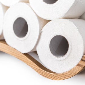 Close-up of a white wall-mounted toilet paper holder with a wave design, holding three rolls of toilet paper. A sleek metal bar supports the rolls, highlighting the wave texture.
