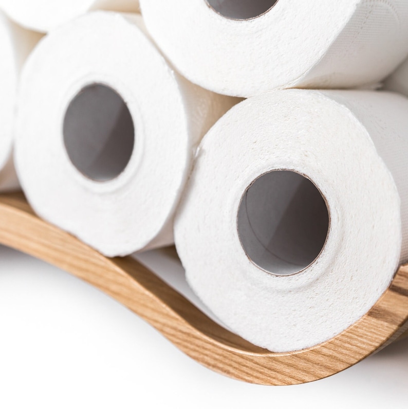 Close-up of a white wall-mounted toilet paper holder with a wave design, holding three rolls of toilet paper. A sleek metal bar supports the rolls, highlighting the wave texture.