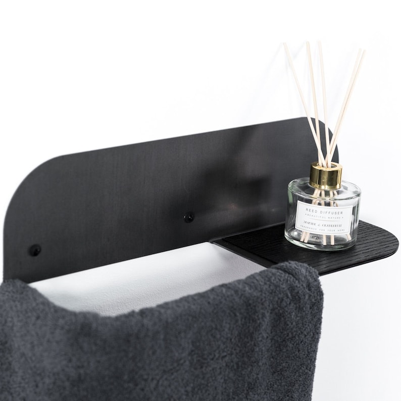 May include: Black metal wall-mounted shelf with a towel bar and a small shelf. A gray towel hangs on the bar and a clear glass bottle with a gold lid and reeds sits on the shelf. The bottle label reads "Reed Diffuser -  Aromatic Nature -  Inspired by grapefruit -  Fragrance for your home".