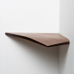 Op de afbeelding: Een zwevend hoekrek van donkerbruin hout. De voorkant is gebogen. Het ontwerp is minimalistisch en modern, geschikt voor het uitstallen van kleine items. De plank is ongeveer 30 cm diep.