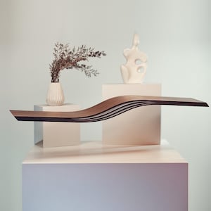 May include: A modern, sculptural wooden shelf with a wave-like design. The shelf is made of layered wood with a dark edge, displayed with a white vase of dried flowers and a white abstract sculpture. The shelf appears to be approximately 60 inches long.