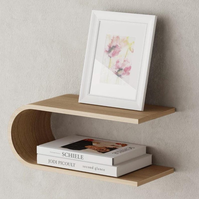 May include: A modern, two-tiered floating shelf made of light-colored wood. A white framed picture with a floral print sits on the top shelf. Two books are stacked on the lower shelf. The shelf has a curved design.