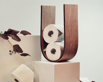 May include: A wooden U-shaped toilet paper holder in a warm brown tone, holding two rolls of white toilet paper. A third roll rests on a white cube, with decorative leaves in the background. The holder is a modern, minimalist design.