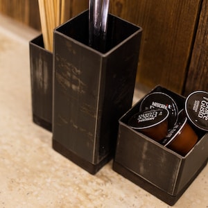 Set of dark metal utensil holders placed on a countertop, one holding ladles, another with wooden skewers, and a smaller holder filled with coffee pods. A wooden wall serves as the background.