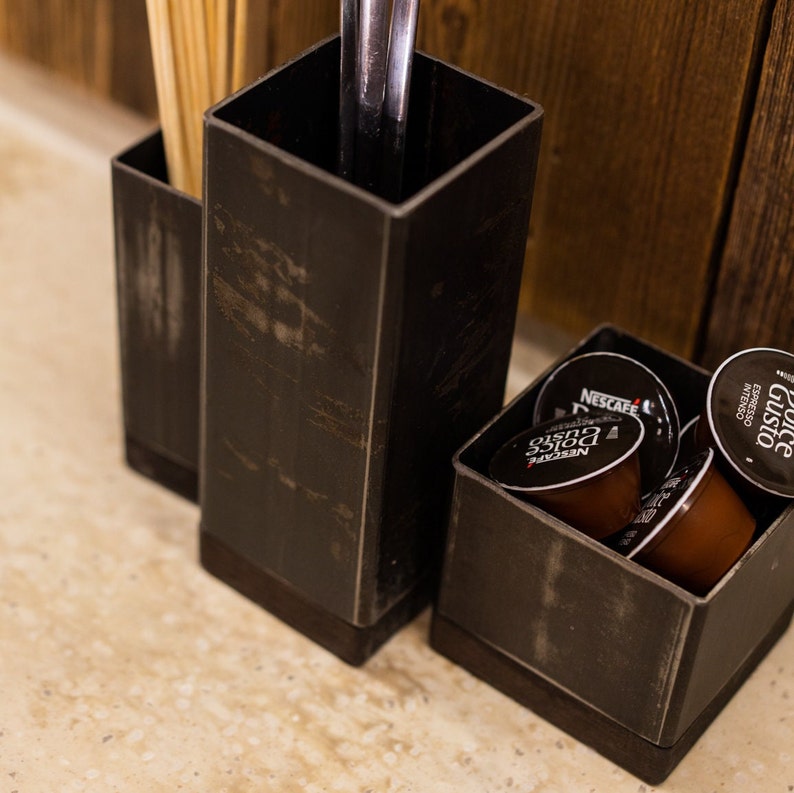 Set of dark metal utensil holders placed on a countertop, one holding ladles, another with wooden skewers, and a smaller holder filled with coffee pods. A wooden wall serves as the background.