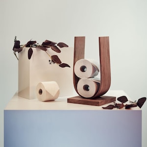 May include: A wooden toilet paper holder with two rolls of toilet paper. The holder is a dark brown, U-shaped design. A single roll of toilet paper sits on a white surface. Decorative branches with brown leaves are also present.