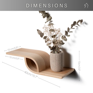 Pu&ograve; includere: Una mensola da parete in legno chiaro con un design curvo unico. La mensola &egrave; lunga 40 cm e alta 10 cm. Un piccolo vaso con fiori secchi &egrave; posto sulla mensola. La parola "DIMENSIONS" &egrave; in alto.
