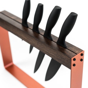 Angled view of a modern knife holder with a dark wooden top, vibrant orange metal frame, and visible black screws, holding four black-handled knives in a single vertical slot.