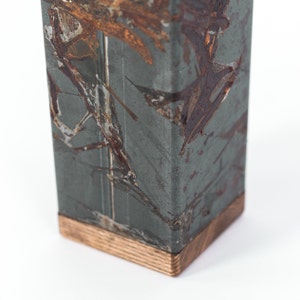 Close-up of a rustic metal utensil holder with a distressed finish, showing a mix of copper and steel tones. The base is made of natural wood, and the holder is displayed against a plain white background.