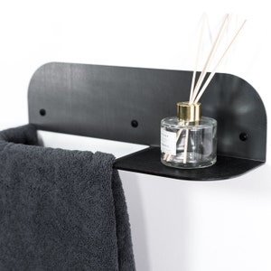May include: A black metal shelf with a curved edge, mounted on a white wall. A gray towel hangs on the left side of the shelf, and a clear glass bottle with a gold lid and wooden sticks sits on the right side of the shelf.